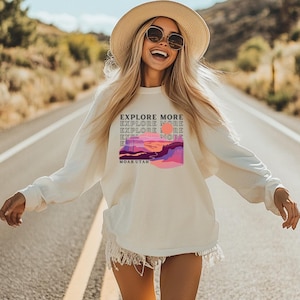 May include: A white long-sleeved sweatshirt with a graphic print of a sunset over a mountain range. The text "EXPLORE MORE" is repeated in a stacked pattern on the graphic. The text "MOAB UTAH" is printed below the graphic.