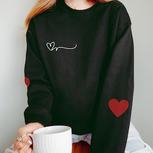 Può includere: Felpa nera con un piccolo disegno a forma di cuore bianco sul petto e cuori rossi sulle maniche.