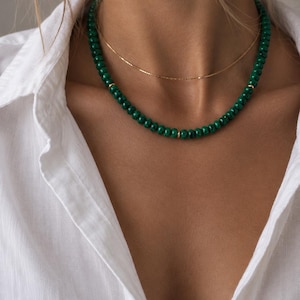 May include: A layered necklace set consisting of a thin gold chain and a beaded necklace featuring small, round, dark green malachite beads interspersed with tiny gold-toned metal spacers, worn over a white collared button-down shirt.