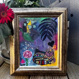 May include: A framed painting of a colorful cat sleeping in a garden setting. The cat is surrounded by flowers, leaves, and a black heart. The background is a vibrant purple and blue.