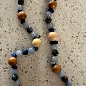 May include: A beaded necklace featuring a variety of natural elements. The necklace includes light blue, dark blue, and brown beads, along with wooden beads and a white seashell pendant. The clasp is gold-colored.