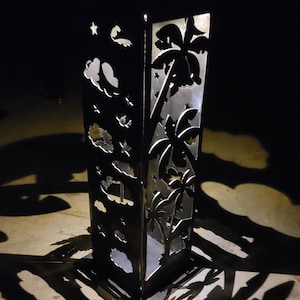 Op de afbeelding: Een hoge, donkere metalen decoratieve pilaar met uitgesneden ontwerpen van palmbomen, wolken en sterren. De pilaar werpt dramatische schaduwen op de grond, waardoor een tropische sfeer ontstaat. Een uniek woondecoratie-item.