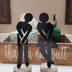 Puede incluir: Dos figuras de metal negro, una masculina y otra femenina, montadas sobre rodajas de madera. Las figuras son siluetas estilizadas. Las figuras están en un entorno de baño, con una bandeja de madera que dice "that's a nut butt".