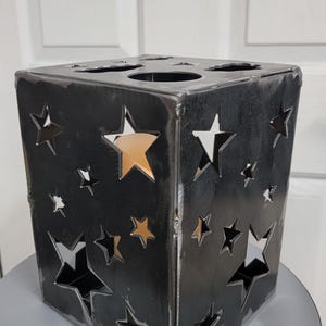 May include: A black metal cube-shaped candle holder with star cutouts. The candle holder has a rustic, industrial look, with visible weld marks. The stars vary in size and are evenly spaced across the sides, allowing light to shine through.