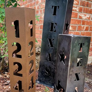 Pode incluir: Três marcadores de endereço de metal. Um é marrom com os números 1224 recortados. Os outros dois são pretos, com a palavra "TEXAS" e uma estrela ou o contorno do Texas recortados. Eles são colocados ao ar livre.