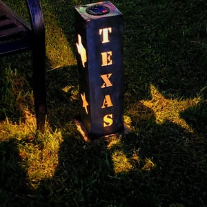 May include: A black solar-powered outdoor light with the word "TEXAS" cut out, along with star and lightning bolt shapes. The light is rectangular and sits on a grassy surface, casting shadows.