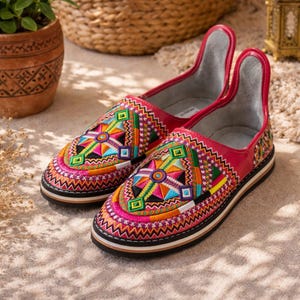 Puede incluir: Un par de zapatos sin cordones bordados con colores vibrantes. Los zapatos presentan una parte superior de cuero rojo con patrones geométricos coloridos en rosa, naranja, verde y azul. Las suelas son negras con una franja blanca y beige. Diseño único.