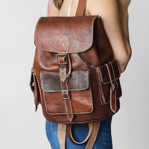 May include: A brown leather backpack with a flap closure, two front pockets, and a side pocket. The backpack has a buckle closure and a strap with a buckle.