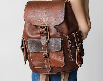 May include: A brown leather backpack with a flap closure, two front pockets, and a side pocket. The backpack has a buckle closure and a strap with a buckle.