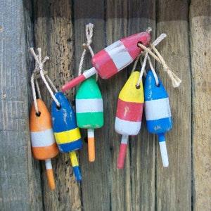 Può includere: Una collezione di sei piccole boe da aragosta in legno dipinte a mano, ognuna con distinte combinazioni di colori tra cui rosso, bianco, blu, verde, giallo e arancione. Ogni boa è attaccata a un breve pezzo di corda naturale per appenderla.