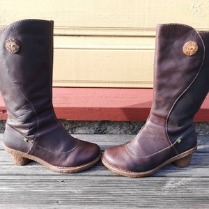 Side profile of El Naturalista boots in dark brown leather with elegant stitching and a rustic cork-textured heel, displayed against a wooden backdrop.