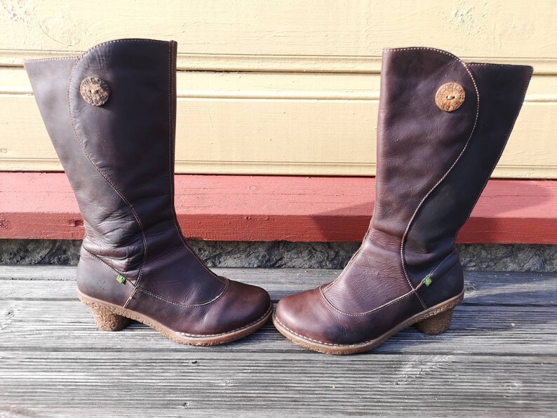 Side profile of El Naturalista boots in dark brown leather with elegant stitching and a rustic cork-textured heel, displayed against a wooden backdrop.
