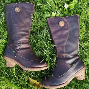 El Naturalista dark brown leather boots laid on lush green grass, showcasing their unique design, nomad style, and cork-textured heels.