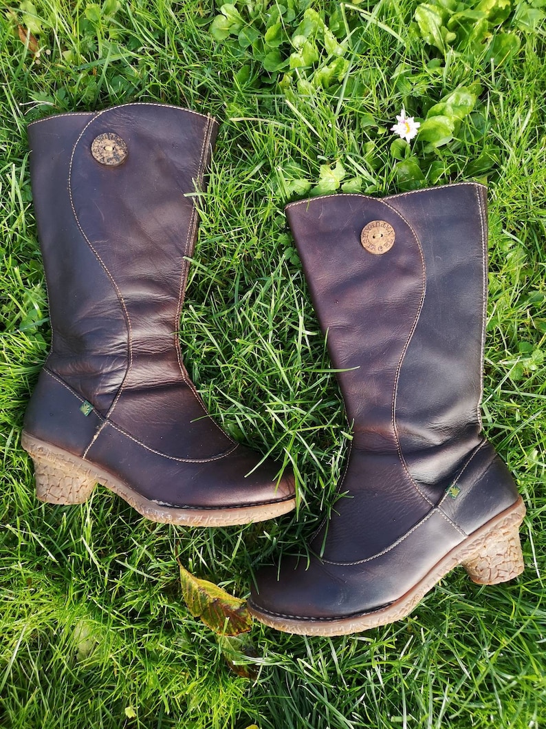 El Naturalista dark brown leather boots laid on lush green grass, showcasing their unique design, nomad style, and cork-textured heels.
