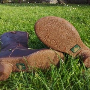 El Naturalista dark brown leather boots lying on green grass, highlighting their unique textured soles and eco-friendly frog logo.