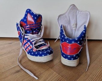 Può includere: Un paio di scarpe da ginnastica alte con un motivo a stelle blu e rosse. Le scarpe hanno una suola bianca e un logo Adidas rosso sul lato.