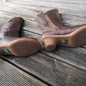 Close-up view of El Naturalista boots on a wooden surface, showcasing the detailed textured soles, short heels, and iconic frog logo.