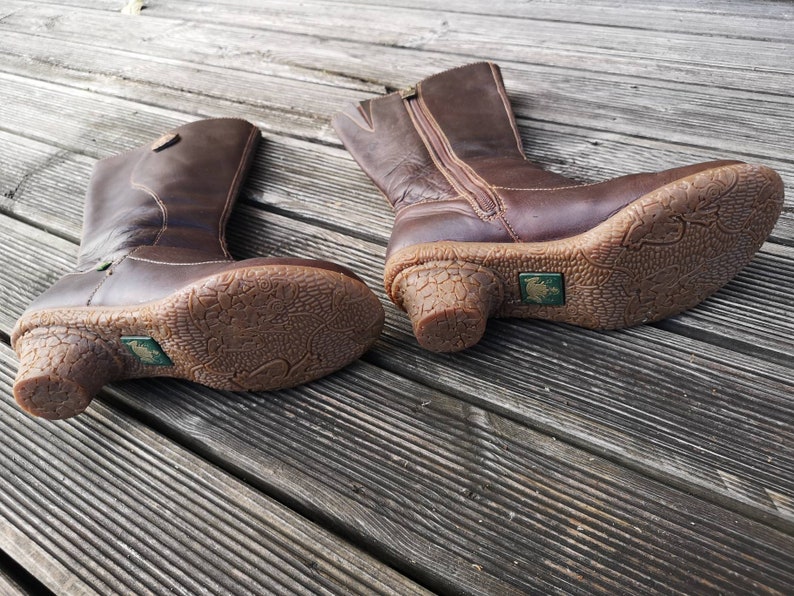 Close-up view of El Naturalista boots on a wooden surface, showcasing the detailed textured soles, short heels, and iconic frog logo.