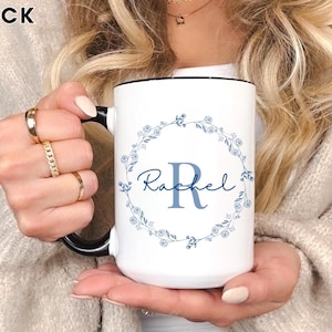 May include: White ceramic mug with a black handle and rim. The mug features a blue floral wreath design with the name "Rachel" and the letter "R" in the center. The word "BLACK" is in the top left corner.