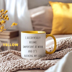 May include: White ceramic mug with a yellow interior and handle. The mug features the text "I ACCIDENTALLY BECAME IMPORTANT AT WORK AND IT'S RUINING MY LIFE" in black lettering. The mug is on a beige knitted blanket.