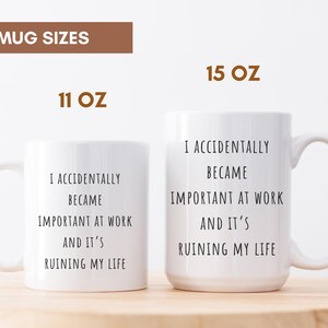 May include: Two white ceramic mugs, 11 oz and 15 oz, with the text "I ACCIDENTALLY BECAME IMPORTANT AT WORK AND IT'S RUINING MY LIFE" in black. The mugs have a curved handle and are on a light brown surface.