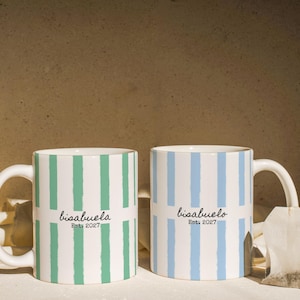 May include: Two white ceramic mugs featuring vertical stripes. The left mug has green stripes and the text "bisabuela Est. 2027". The right mug has light blue stripes and the text "bisabuelo Est. 2027". A tea bag sits next to the right mug.