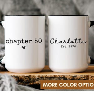 May include: Two white ceramic mugs with black handles and rims. The left mug features the text "chapter 50" with a small heart icon. The right mug displays the name "Charlotte" in script font above the text "Est. 1976" in a typewriter font.