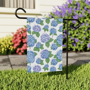 May include: A garden flag featuring a pattern of blue and purple hydrangea flowers with green leaves on a white background. The text "fuck trump" is repeated throughout the design. The flag is displayed on a black metal stand.