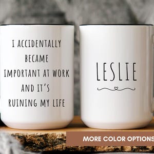 May include: Two white ceramic mugs with black handles and rims. One mug reads "I ACCIDENTALLY BECAME IMPORTANT AT WORK AND IT'S RUINING MY LIFE." The other mug has the name "LESLIE" with a heart design. The mugs are on a wooden surface.