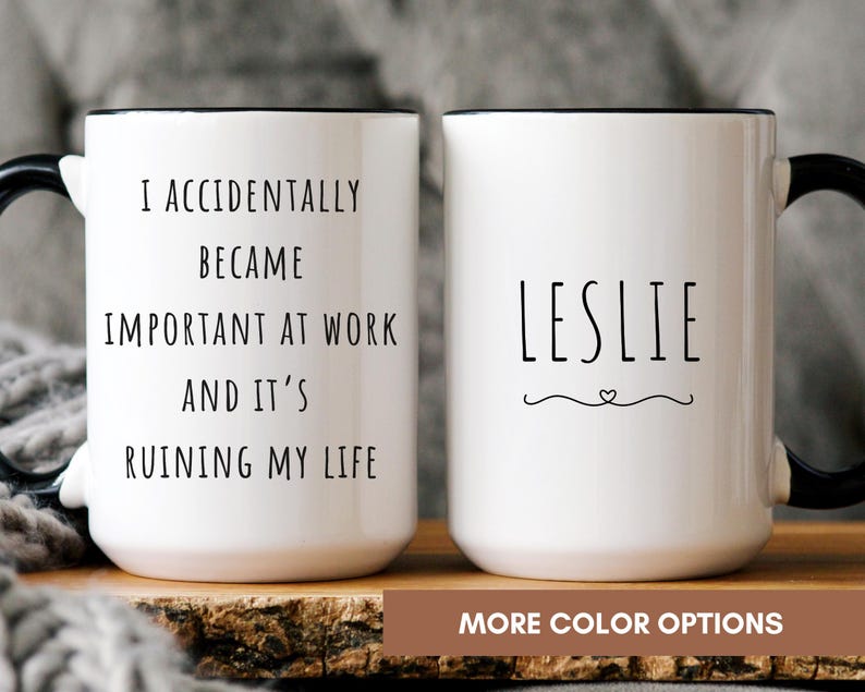 May include: Two white ceramic mugs with black handles and rims. One mug reads "I ACCIDENTALLY BECAME IMPORTANT AT WORK AND IT'S RUINING MY LIFE." The other mug has the name "LESLIE" with a heart design. The mugs are on a wooden surface.