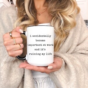 May include: White ceramic mug with a black handle and rim. The mug features the text "I accidentally became important at work and it's ruining my life" in black lettering. The mug is held by a person.