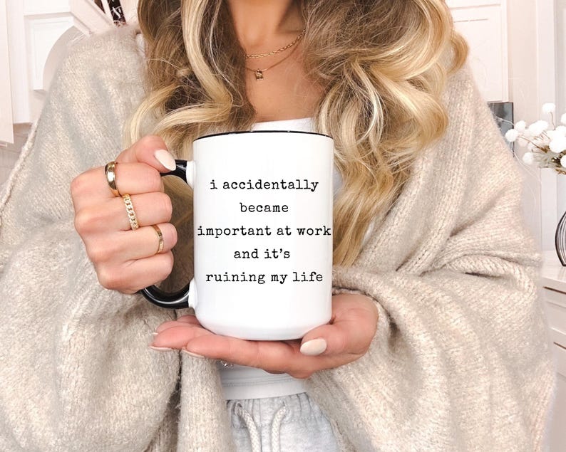 May include: White ceramic mug with a black handle and rim. The mug features the text "I accidentally became important at work and it's ruining my life" in black lettering. The mug is held by a person.