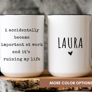 May include: Two white ceramic mugs with black handles and rims. One mug has the text "i accidentally became important at work and it's ruining my life." The other mug has the name "LAURA" and a heart symbol. The mugs are on a wooden surface.