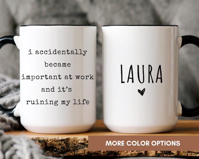 May include: Two white ceramic mugs with black handles and rims. One mug has the text "i accidentally became important at work and it's ruining my life." The other mug has the name "LAURA" and a heart symbol. The mugs are on a wooden surface.