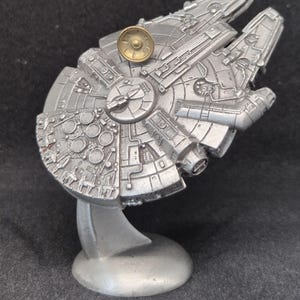 May include: A silver model of the Millennium Falcon spaceship from Star Wars, on a silver stand. The model has a gold-coloured disc on top.