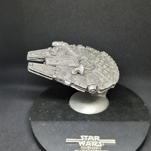Może przedstawiać: Srebrny model statku kosmicznego Millennium Falcon z Gwiezdnych Wojen. Model stoi na czarnej podstawie z napisem "Star Wars" i "Han Solo's Millennium Falcon".