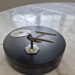 Puede incluir: Un modelo plateado de la Starship Enterprise, una nave espacial de la franquicia Star Trek. El modelo está montado sobre una base negra y circular. La nave presenta una sección de platillo, dos góndolas de curvatura y un casco secundario.