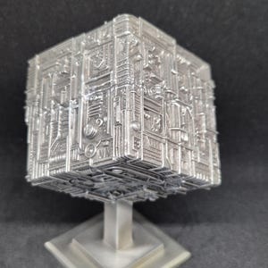 May include: A silver metal sculpture of a cube with intricate details, mounted on a square base with a pillar.