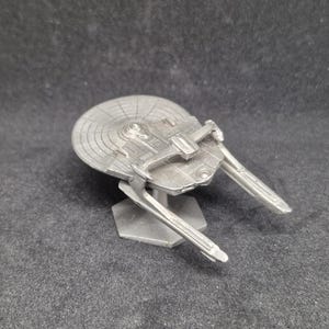 Puede incluir: Modelo plateado de una nave espacial, con un casco principal circular y dos largas góndolas de curvatura. El modelo está montado sobre una base hexagonal, y los detalles de la nave están finamente grabados.