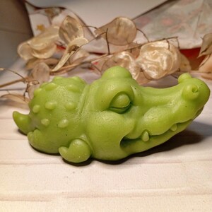 May include: A light green alligator-shaped soap. The soap has a textured surface with a detailed design of the alligator's head, body, and legs. The soap is resting on a white surface.