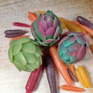 Puede incluir: Tres jabones con forma de alcachofa en verde, rosa y turquesa, rodeados de jabones con forma de zanahoria de colores rojo, naranja, morado y amarillo. Los jabones están dispuestos sobre una superficie de madera clara.
