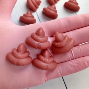 May include: Several small, reddish-brown soaps shaped like a pile of poop. The soaps are arranged on a pink surface, with others scattered in the background. The soaps have a detailed, textured design.