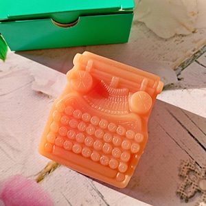 Könnte beinhalten: Pfirsichfarbene Seife in Form einer alten Schreibmaschine. Die Seife hat detaillierte Tasten und runde Knöpfe. Eine grüne Schachtel ist teilweise im Hintergrund zu sehen. Die Seife liegt auf einer weißen und hellbraunen Oberfläche.