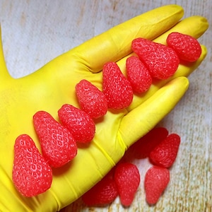 May include: A yellow gloved hand holds a row of small, bright red, strawberry-shaped sweets. More sweets are scattered on a wooden surface.