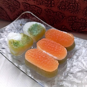 Pode incluir: Um prato de vidro transparente contém vários sabonetes em forma de sushi. Dois sabonetes triangulares têm arroz branco e detalhes em algas verdes. Três sabonetes retangulares têm tampas laranja e bases de arroz branco.