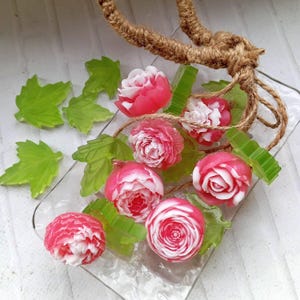 May include: Handmade soaps in the shape of pink and white roses and green leaves, presented on a clear glass plate. The soaps are detailed, with a natural rope accent.