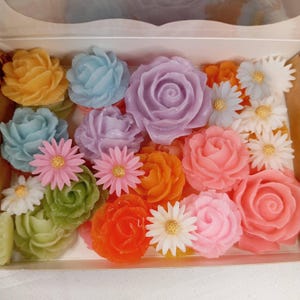 Könnte beinhalten: Eine Schachtel voller farbenfroher, handgefertigter Blumenarrangements. Das Sortiment umfasst Rosen, Gänseblümchen und andere Blütenformen in Gelb-, Blau-, Lila-, Orange-, Grün- und Rosatönen. Die Blumen sind in einer weißen Schachtel angeordnet.