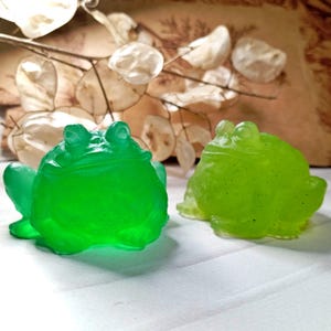May include: Two frog-shaped figurines in shades of green. One is a vibrant, translucent green, and the other is a lighter, semi-transparent green. The frogs are detailed with visible eyes and textured skin, suggesting a whimsical or decorative theme.