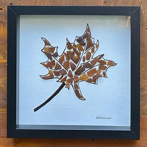 May include: Framed artwork featuring a maple leaf design. The leaf is composed of brown and amber-colored glass pieces, outlined in black, and set against a white background. The artwork is encased in a black frame.
