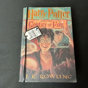 May include: A hardcover book titled Harry Potter and the Goblet of Fire by J.K. Rowling with a teal and orange cover illustration featuring a character with glasses holding a wand. A black elastic band holds a tag labeled Secret Compartment Books.